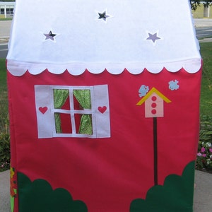 May include: A red play tent with a white roof and scalloped edges. The tent has a window with green and white curtains, a birdhouse with red dots, and two green bushes on the bottom.