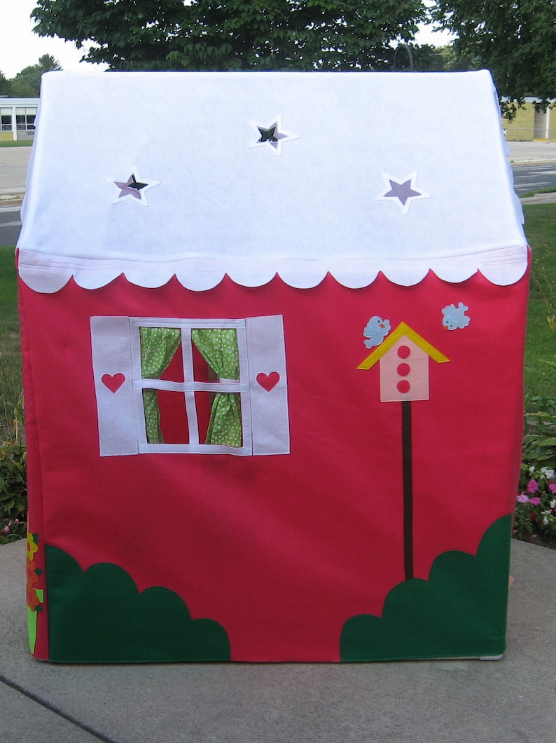 May include: A red play tent with a white roof and scalloped edges. The tent has a window with green and white curtains, a birdhouse with red dots, and two green bushes on the bottom.