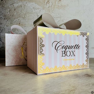 Puede incluir: Una caja de rayas rosa y blancas con ribetes dorados y las palabras "Coquette Box" en una fuente dorada. La caja está atada con una cinta blanca.