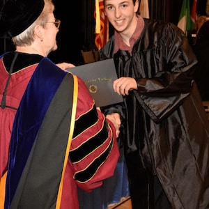 May include: A person in a black graduation gown and cap is shaking hands with a person wearing a red and gold academic gown. The person in the gown is holding a diploma that says "Baylor College".