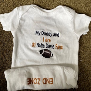 May include: White baby onesie with the text "My Daddy and I are #1 Notre Dame Fans" in navy blue and orange embroidery. A brown football is embroidered below the text. The onesie has the text "END ZONE" in orange embroidery on the bottom.