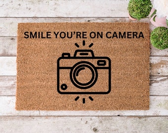 Może przedstawiać: Brązowa mata kokosowa z napisem "SMILE YOU'RE ON CAMERA" w czarnych literach. Czarno-biały zarys aparatu z lampą błyskową jest wyśrodkowany na macie.