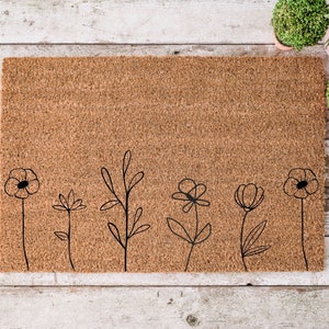 May include: A brown coir doormat with a simple design of five black flowers and stems. The doormat is on a white wooden surface.