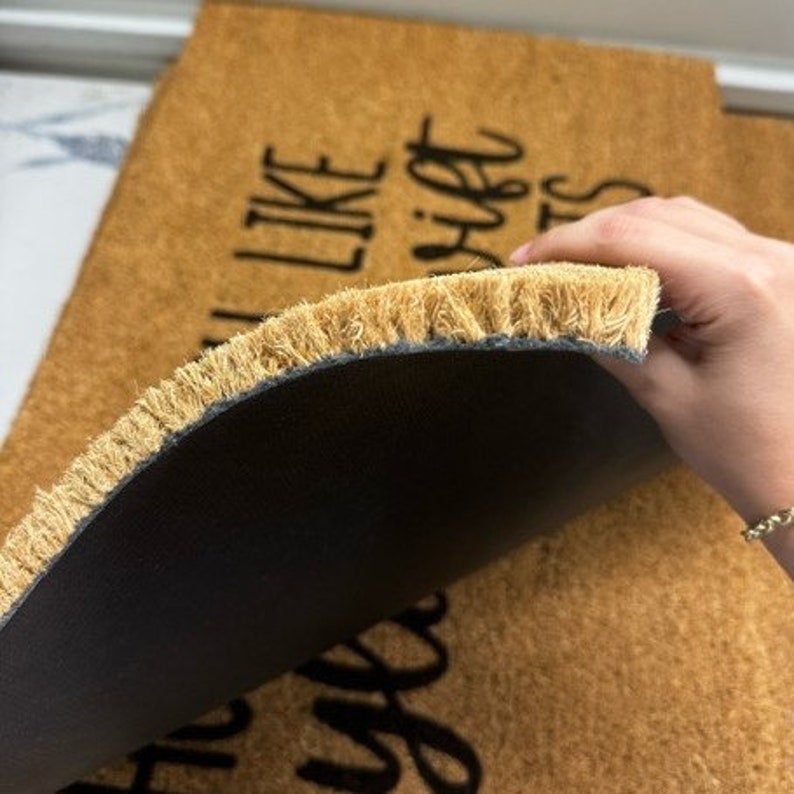 Op de afbeelding: Een bruine kokosvezel deurmat met de tekst "I'm all like" en "high five" in het zwart. De achterkant van de deurmat is zwart en heeft een bruine franje.