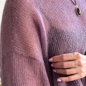 May include: A sheer, lavender-colored sweater with a loose weave. The sweater has a boat neck and long sleeves. A dark pendant hangs from a thin cord. The image shows a close-up of the sweater's texture and the wearer's hand.