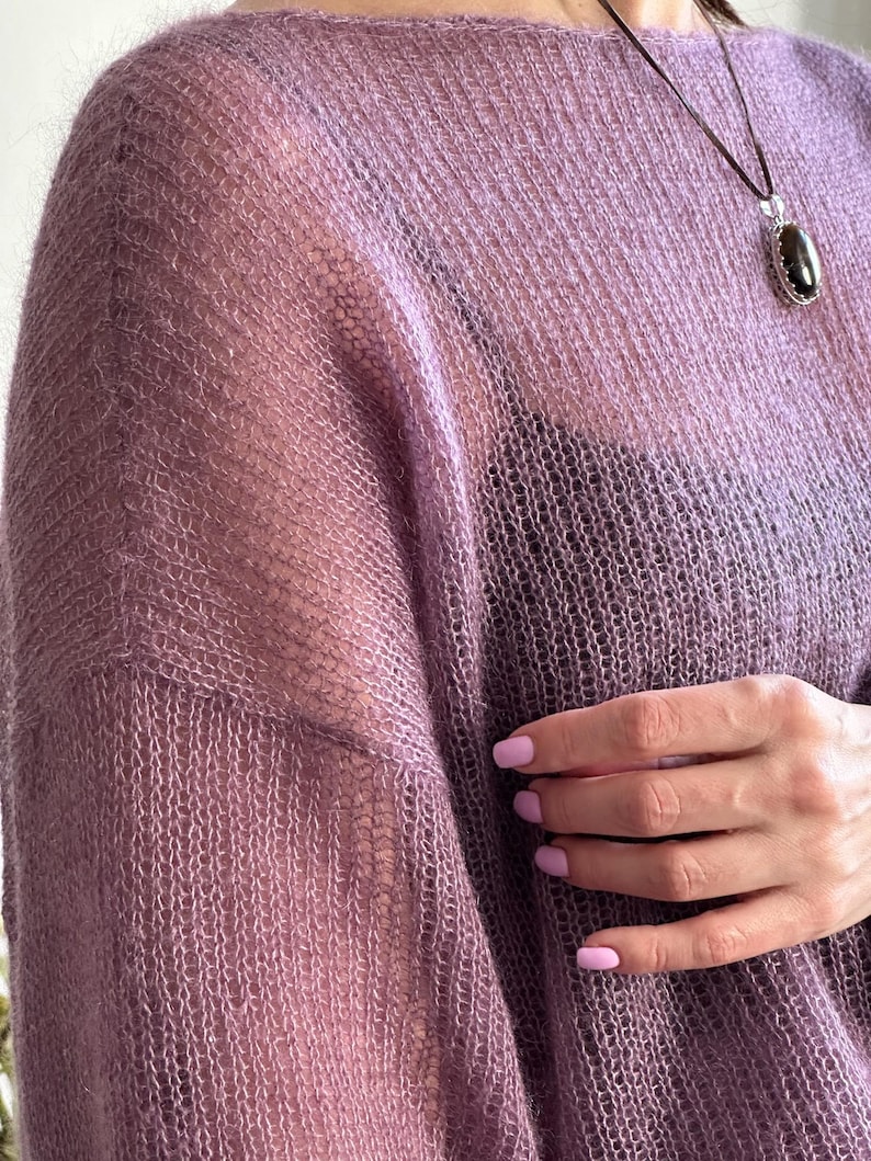 May include: A sheer, lavender-colored sweater with a loose weave. The sweater has a boat neck and long sleeves. A dark pendant hangs from a thin cord. The image shows a close-up of the sweater's texture and the wearer's hand.
