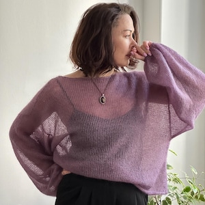 May include: A sheer, lavender-colored sweater with long, voluminous sleeves. The sweater has a wide neckline and is paired with black pants. A pendant necklace is visible. The overall style is casual and airy.