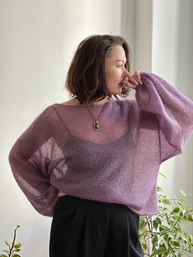 May include: A sheer, lavender-colored sweater with long, voluminous sleeves. The sweater has a wide neckline and is paired with black pants. A pendant necklace is visible. The overall style is casual and airy.