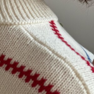 May include: Close-up of a cream-colored sweater with red and blue embroidered details. The sweater features a ribbed neckline and red diagonal stitching across the front. The knitwear appears soft and comfortable.