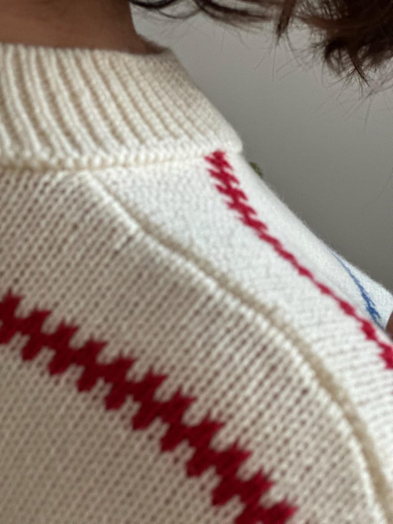 May include: Close-up of a cream-colored sweater with red and blue embroidered details. The sweater features a ribbed neckline and red diagonal stitching across the front. The knitwear appears soft and comfortable.