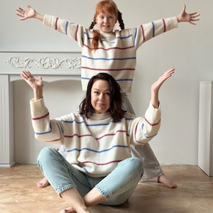May include: Two people wearing matching cream-colored sweaters with horizontal red and blue stripes. The sweaters have long sleeves and a crew neck. The person in the foreground is wearing light blue jeans. The background is a plain white wall.