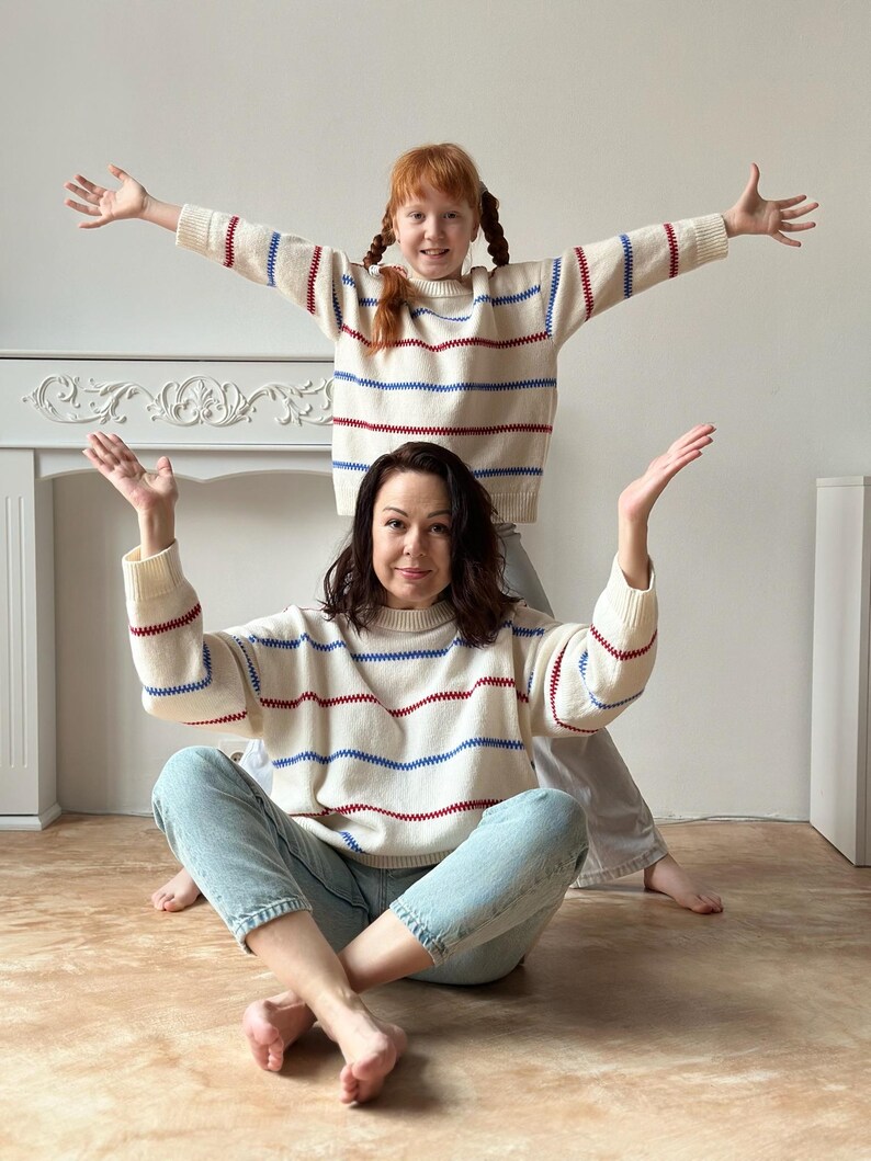 May include: Two people wearing matching cream-colored sweaters with horizontal red and blue stripes. The sweaters have long sleeves and a crew neck. The person in the foreground is wearing light blue jeans. The background is a plain white wall.