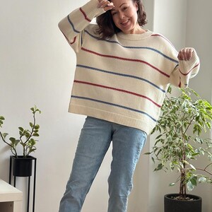 May include: Cream-colored sweater with horizontal red and blue stripes. The sweater has a round neckline and long sleeves. The person is wearing light blue jeans. The sweater is a casual, everyday garment.