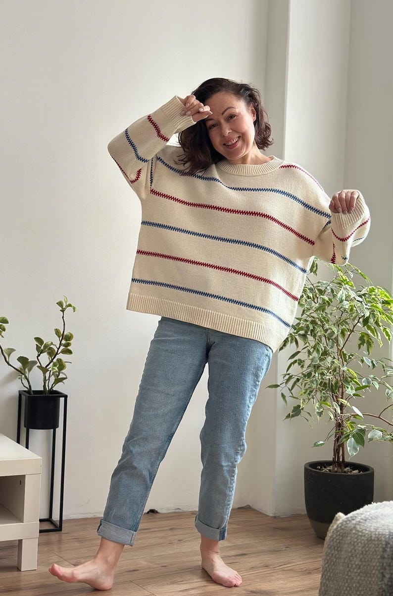 May include: Cream-colored sweater with horizontal red and blue stripes. The sweater has a round neckline and long sleeves. The person is wearing light blue jeans. The sweater is a casual, everyday garment.