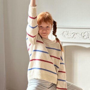 May include: A cream-colored sweater with horizontal red and blue stripes. The sweater is worn by a person with red hair and light gray pants. The person has their arm raised in a fist.