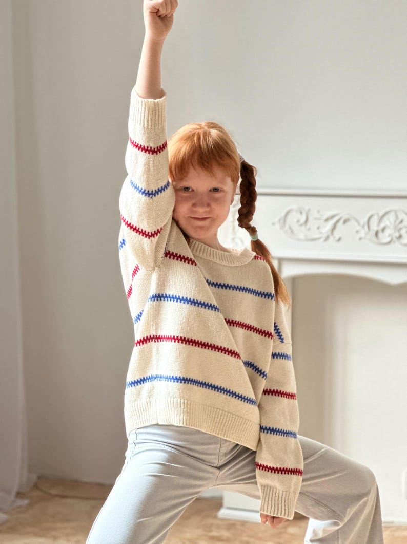 May include: A cream-colored sweater with horizontal red and blue stripes. The sweater is worn by a person with red hair and light gray pants. The person has their arm raised in a fist.