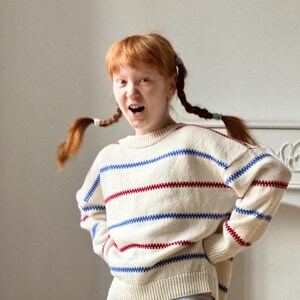 May include: Cream-colored sweater with horizontal blue and red stripes. The sweater has a crew neck and long sleeves. The stripes are a zig-zag pattern. The sweater is worn by a person with red hair in pigtails.