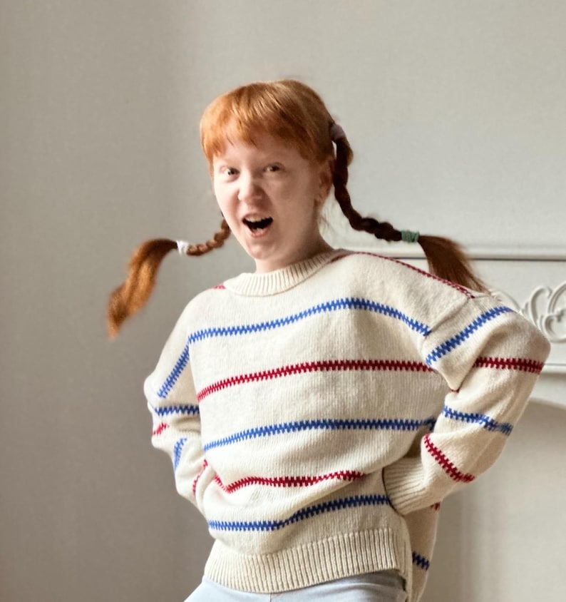 May include: Cream-colored sweater with horizontal blue and red stripes. The sweater has a crew neck and long sleeves. The stripes are a zig-zag pattern. The sweater is worn by a person with red hair in pigtails.