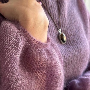 May include: A lavender-colored, open-weave sweater with long sleeves. A brown cord necklace with a pendant featuring a dark stone and silver accents hangs over the sweater. The sweater's texture is soft and fuzzy.