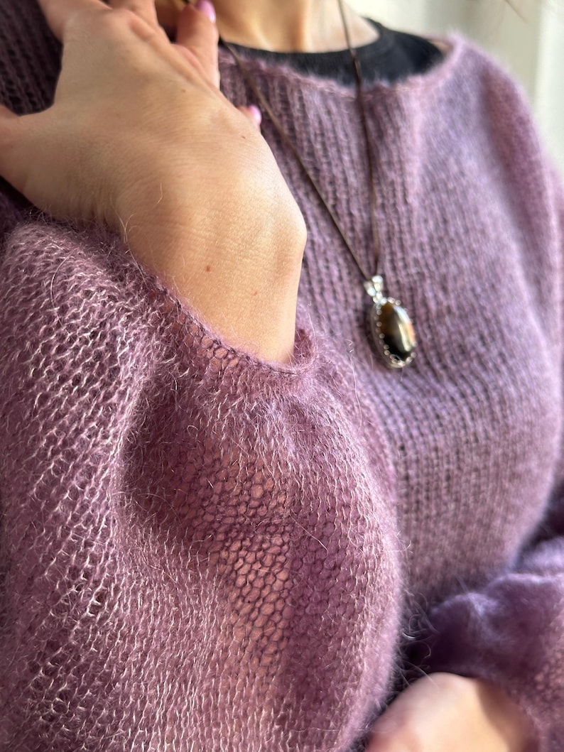 May include: A lavender-colored, open-weave sweater with long sleeves. A brown cord necklace with a pendant featuring a dark stone and silver accents hangs over the sweater. The sweater's texture is soft and fuzzy.