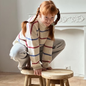 May include: A young person with red hair in pigtails wearing a cream-colored sweater with red and blue stripes, and gray pants, crouches on two small wooden stools. The stools have a round seat and four legs.