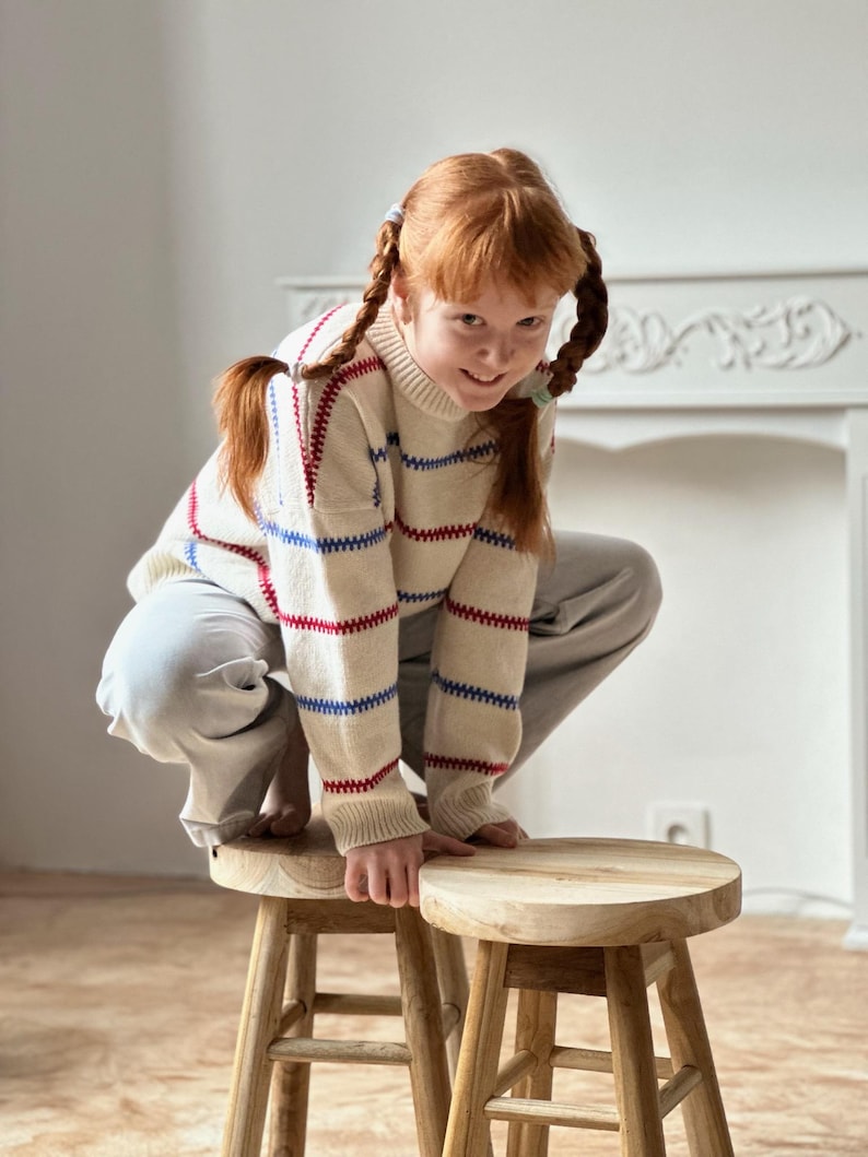 May include: A young person with red hair in pigtails wearing a cream-colored sweater with red and blue stripes, and gray pants, crouches on two small wooden stools. The stools have a round seat and four legs.