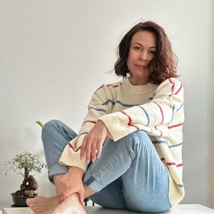 May include: Cream-colored sweater with red and blue striped accents. The sweater has a crew neck and long sleeves. The person is wearing light blue jeans. The overall look is casual and comfortable.