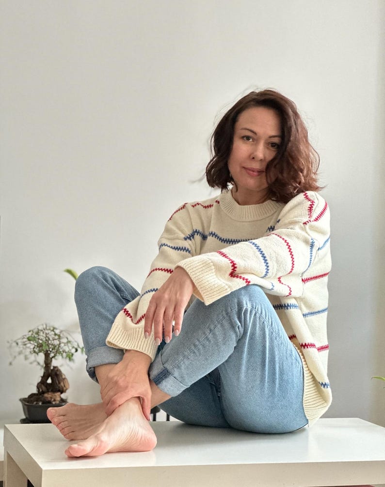 May include: Cream-colored sweater with red and blue striped accents. The sweater has a crew neck and long sleeves. The person is wearing light blue jeans. The overall look is casual and comfortable.