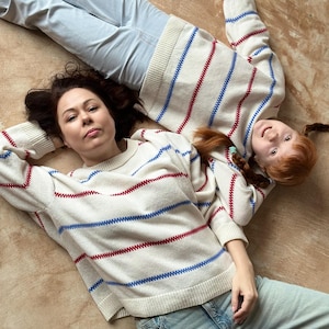 May include: Two people wearing matching cream-colored sweaters with red and blue stripes, paired with light blue jeans, are lying on a brown surface. The sweaters have a classic, casual design, suitable for everyday wear. The overall aesthetic is relaxed and comfortable.