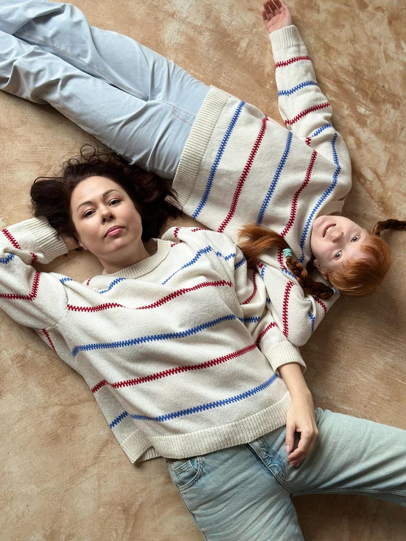 May include: Two people wearing matching cream-colored sweaters with red and blue stripes, paired with light blue jeans, are lying on a brown surface. The sweaters have a classic, casual design, suitable for everyday wear. The overall aesthetic is relaxed and comfortable.