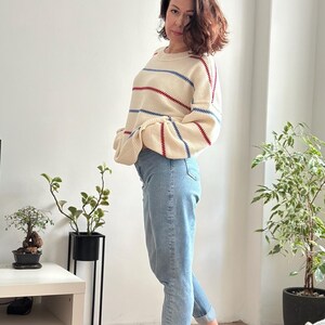 May include: A cream-colored sweater with horizontal red and blue stripes is paired with light blue jeans. The sweater has a round neckline and long sleeves. The jeans are cuffed at the ankles.
