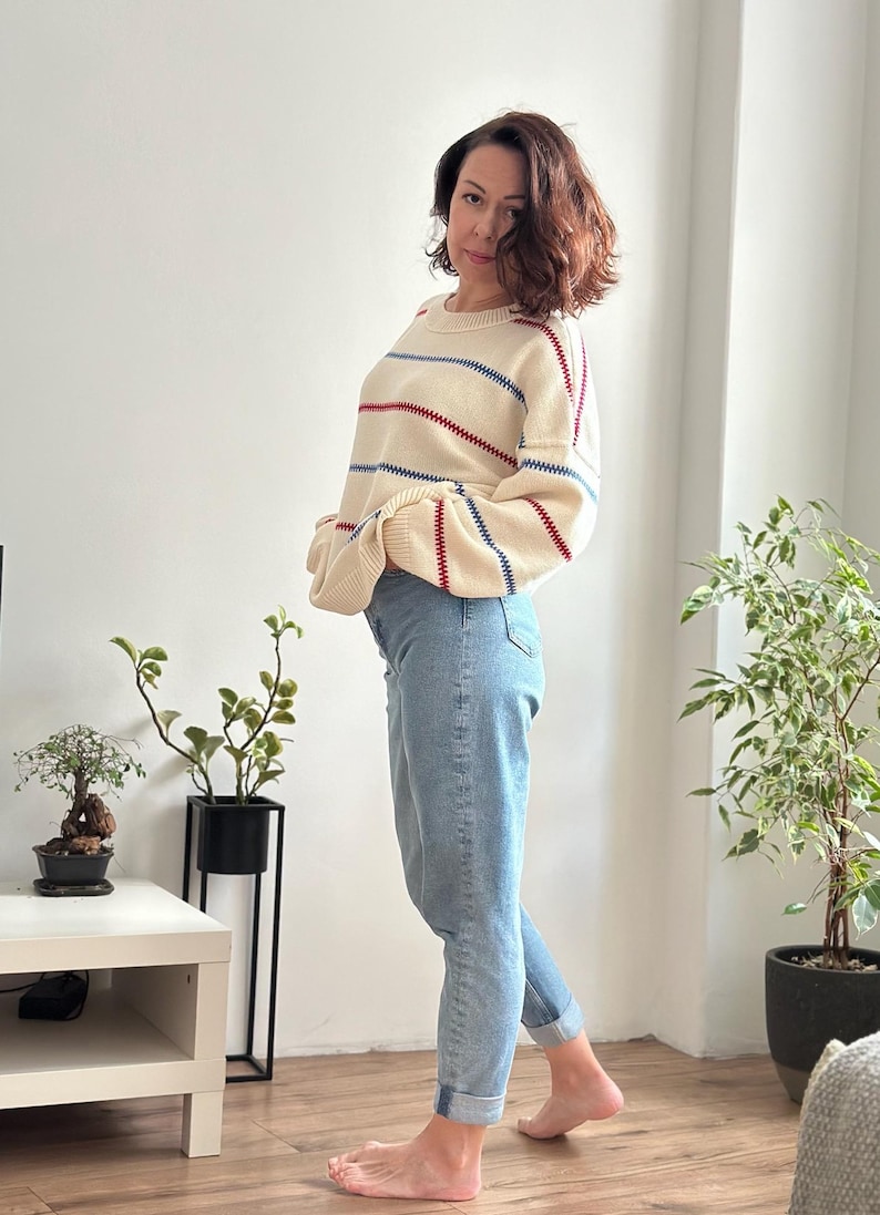 May include: A cream-colored sweater with horizontal red and blue stripes is paired with light blue jeans. The sweater has a round neckline and long sleeves. The jeans are cuffed at the ankles.
