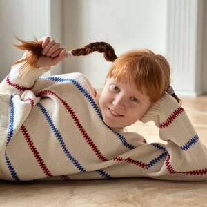 May include: A cream-colored sweater with red and blue stripes. The sweater has a turtleneck and long sleeves. The person in the image is holding their red hair braids. The sweater is a cozy and comfortable garment.