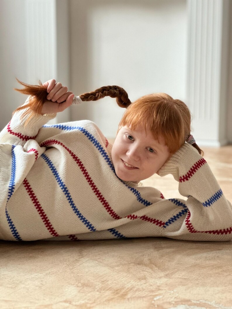 May include: A cream-colored sweater with red and blue stripes. The sweater has a turtleneck and long sleeves. The person in the image is holding their red hair braids. The sweater is a cozy and comfortable garment.
