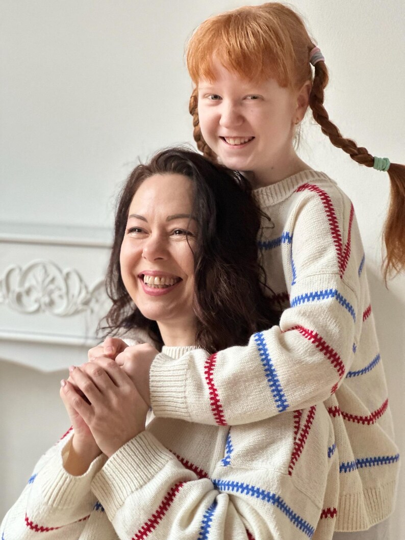 May include: Two people wearing matching cream-colored sweaters with red and blue striped accents. The sweaters have a classic, comfortable design. The people are smiling, creating a warm and inviting image.
