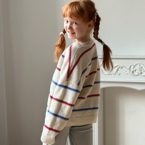 May include: A cream-colored sweater with red and blue horizontal stripes. The sweater is worn by a person with red hair in pigtails, wearing gray pants. The person is smiling and looking over their shoulder.