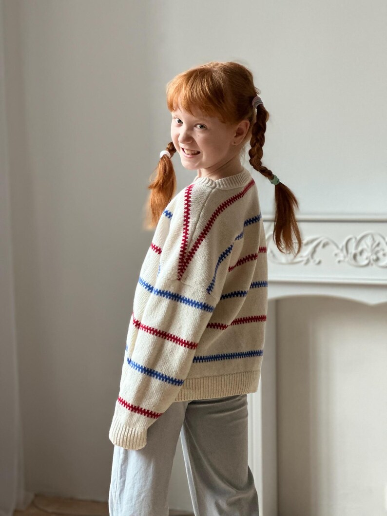 May include: A cream-colored sweater with red and blue horizontal stripes. The sweater is worn by a person with red hair in pigtails, wearing gray pants. The person is smiling and looking over their shoulder.