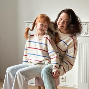 May include: Two people wearing matching cream-colored sweaters with red and blue horizontal stripes. The sweaters are paired with light-wash jeans. One person is sitting on a small stool, and the other is standing behind them.