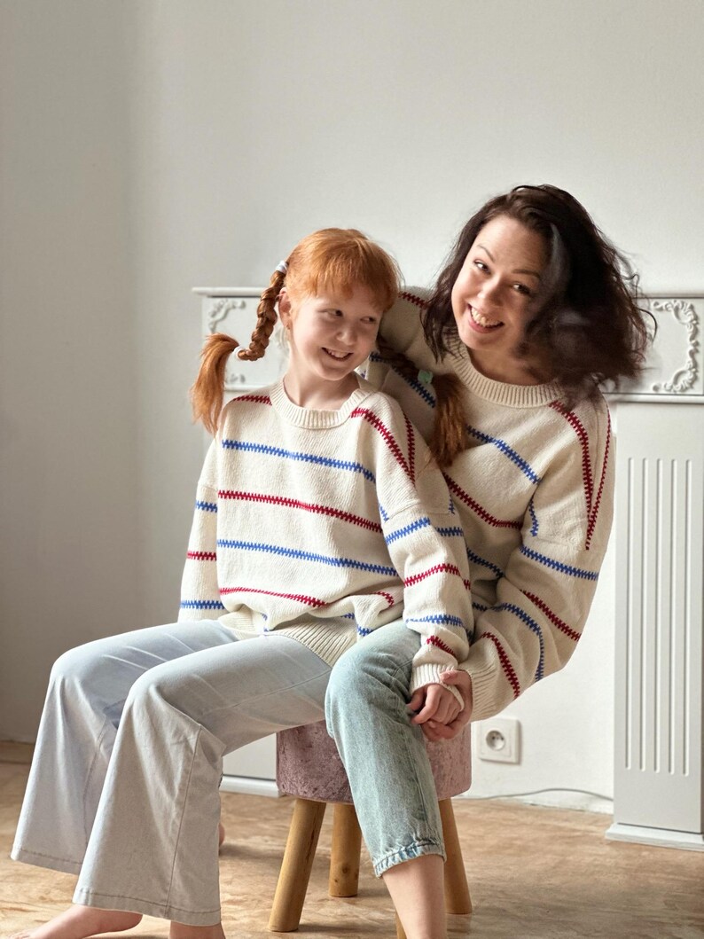 May include: Two people wearing matching cream-colored sweaters with red and blue horizontal stripes. The sweaters are paired with light-wash jeans. One person is sitting on a small stool, and the other is standing behind them.