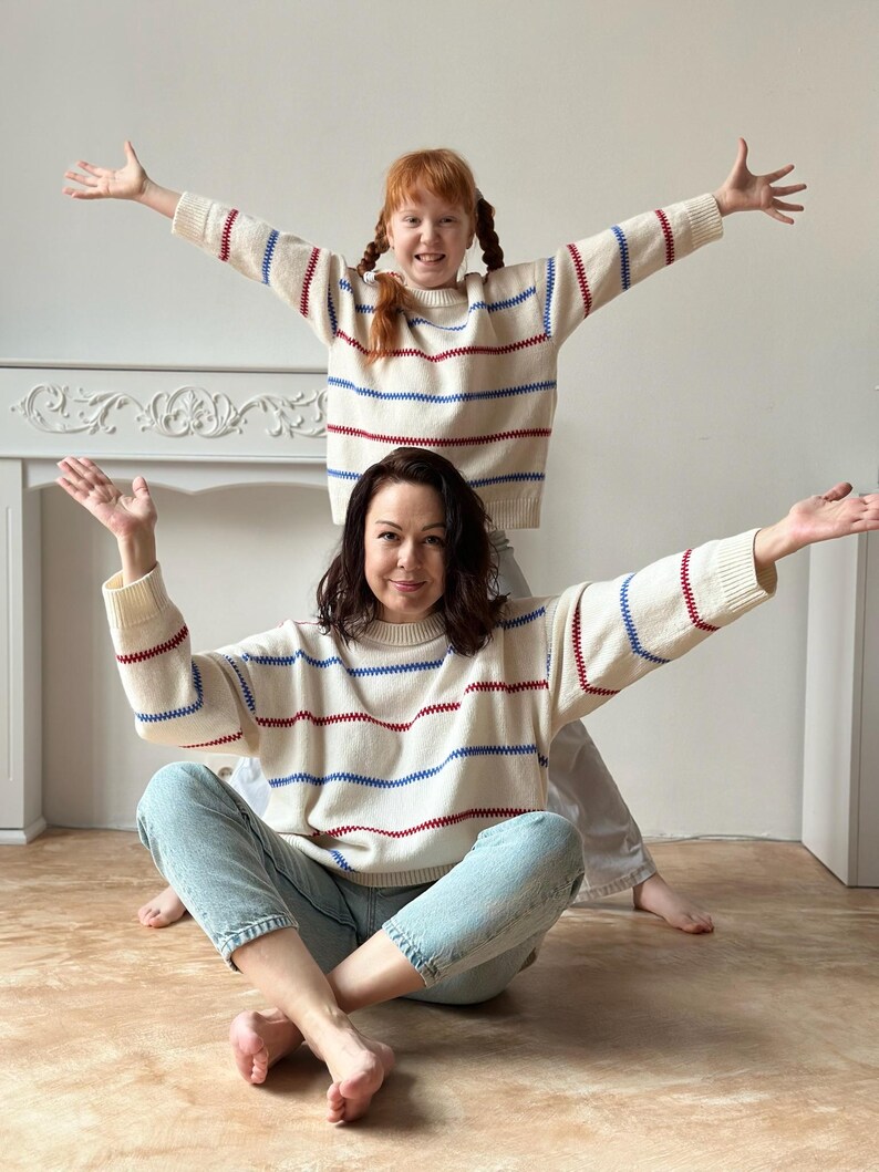 May include: Two people wearing matching cream-colored sweaters with horizontal red and blue stripes. The sweaters have long sleeves and a crew neck. The person in the foreground is sitting cross-legged, while the other person is standing behind them.