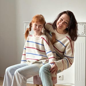 May include: Two people wearing matching cream-colored sweaters with red and blue horizontal stripes. The sweaters are paired with light-wash jeans. The people are sitting on a small stool in front of a white wall.