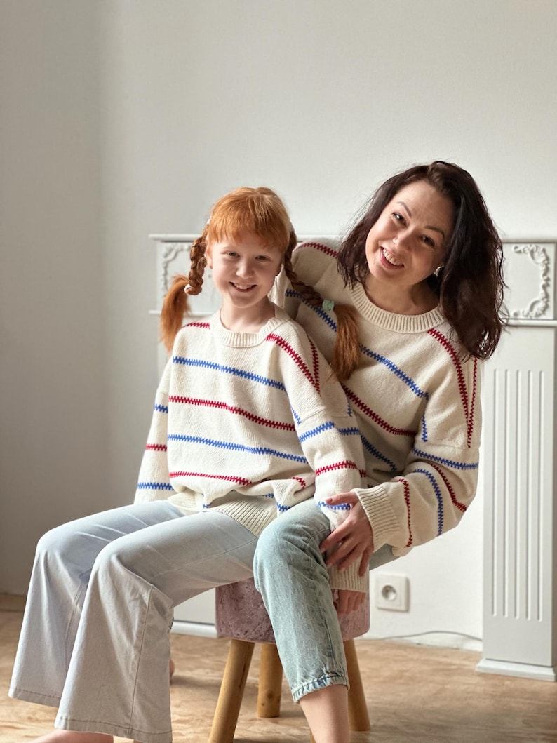 May include: Two people wearing matching cream-colored sweaters with red and blue horizontal stripes. The sweaters are paired with light-wash jeans. The people are sitting on a small stool in front of a white wall.