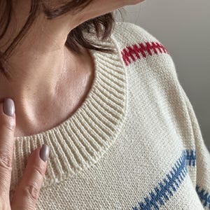May include: Cream-colored sweater with a ribbed crew neck and red and blue striped accents. The sweater has a classic design, with the stripes across the chest and shoulders. The knitwear is a comfortable, casual style.