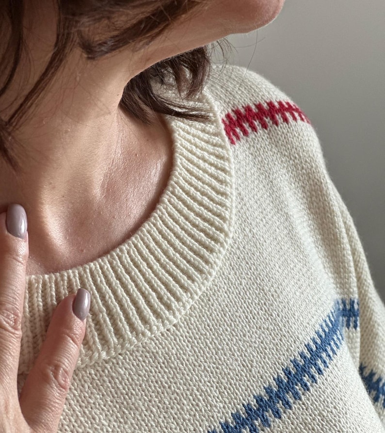 May include: Cream-colored sweater with a ribbed crew neck and red and blue striped accents. The sweater has a classic design, with the stripes across the chest and shoulders. The knitwear is a comfortable, casual style.