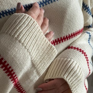 May include: Cream-colored knit sweater with horizontal stripes in red and blue. The sweater has ribbed cuffs and a crew neck. The sleeves are long and the knit is a medium weight. The sweater is a classic style.