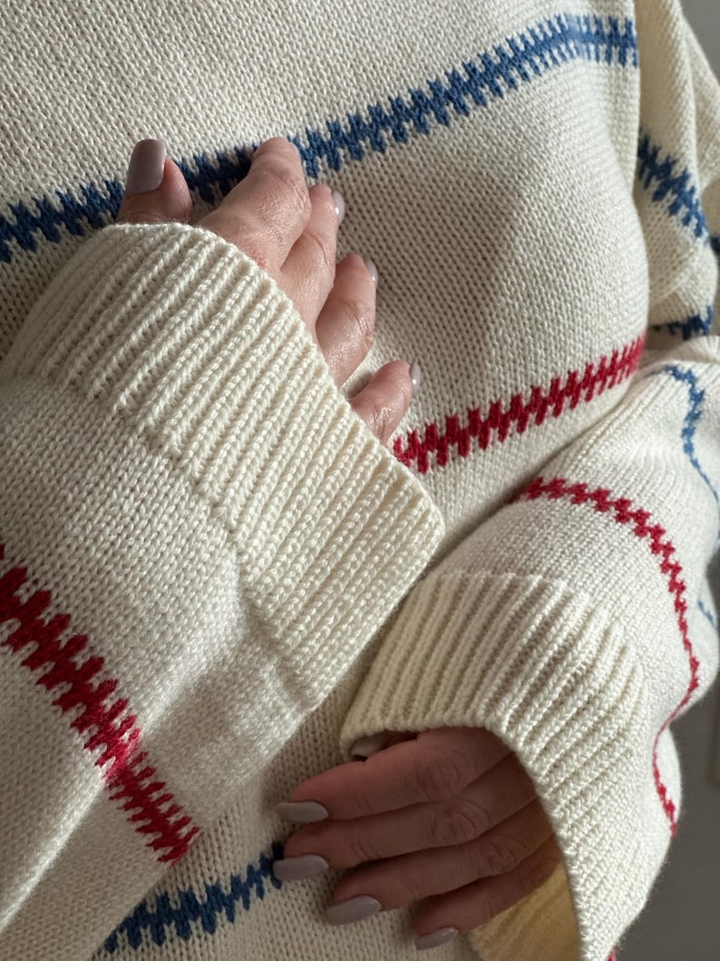 May include: Cream-colored knit sweater with horizontal stripes in red and blue. The sweater has ribbed cuffs and a crew neck. The sleeves are long and the knit is a medium weight. The sweater is a classic style.