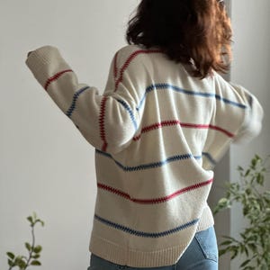 May include: Cream-colored sweater with horizontal red and blue stripes. The sweater has a relaxed fit and long sleeves. The stitching is visible, adding a textured detail. The person is wearing blue jeans.