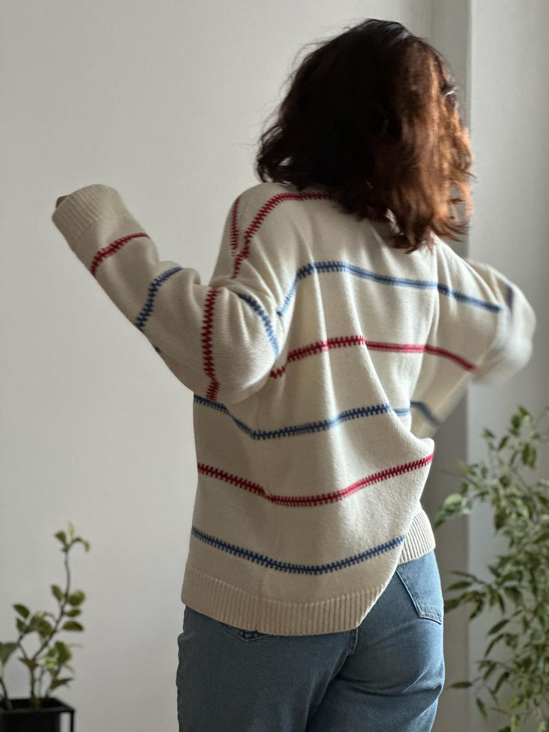 May include: Cream-colored sweater with horizontal red and blue stripes. The sweater has a relaxed fit and long sleeves. The stitching is visible, adding a textured detail. The person is wearing blue jeans.