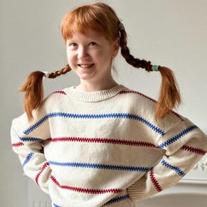 May include: A cream-colored sweater with horizontal red and blue stripes. The sweater has a round neckline and long sleeves. The stripes are evenly spaced across the chest and arms. The sweater is made of a soft, knitted material.
