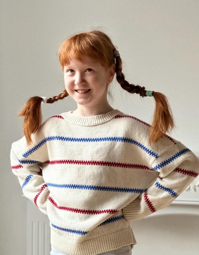 May include: A cream-colored sweater with horizontal red and blue stripes. The sweater has a round neckline and long sleeves. The stripes are evenly spaced across the chest and arms. The sweater is made of a soft, knitted material.